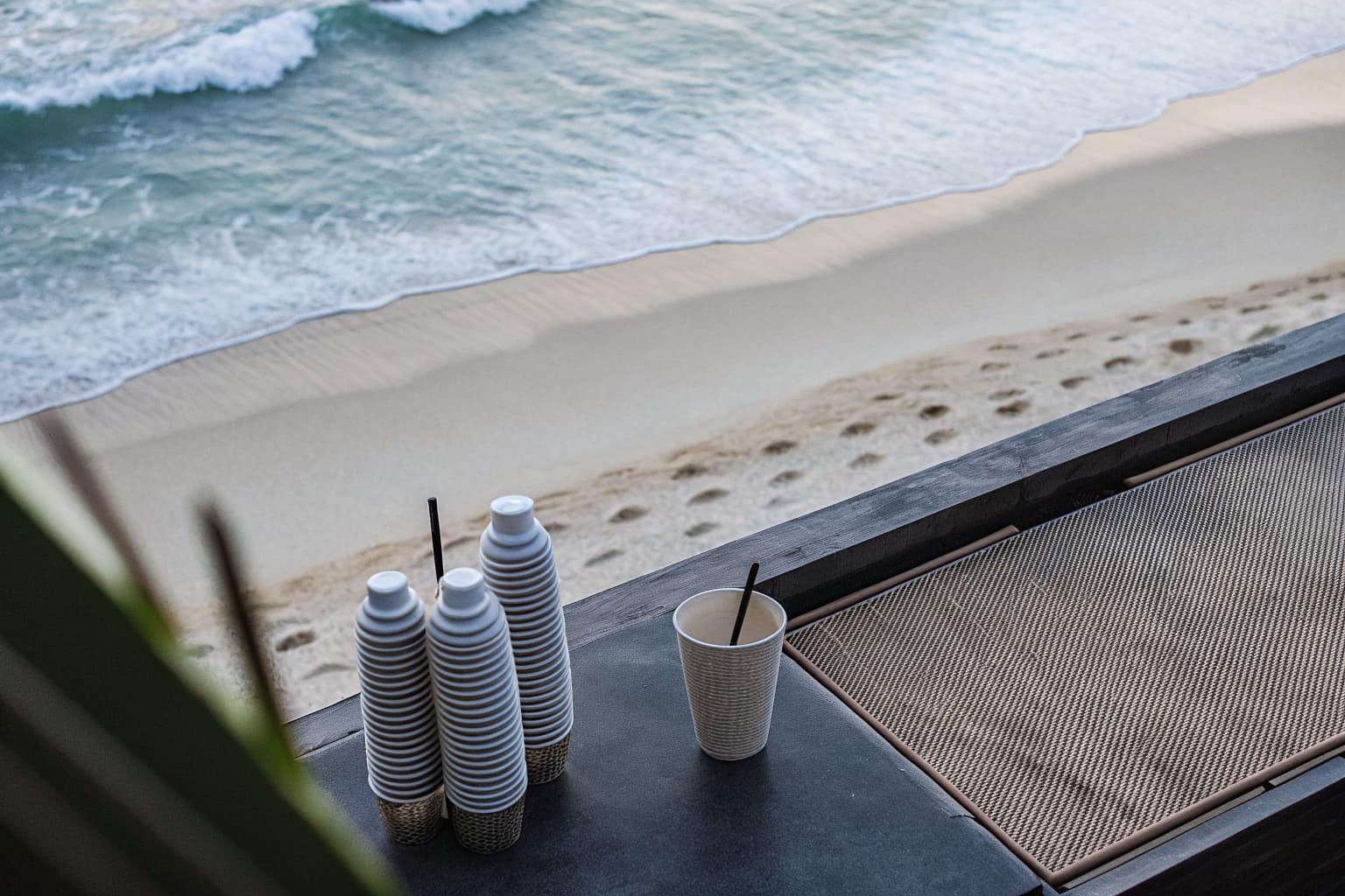 A resort pool deck with biodegradable PHA cups on a table beside an ocean shoreline where discarded plastic waste rests in the sand
